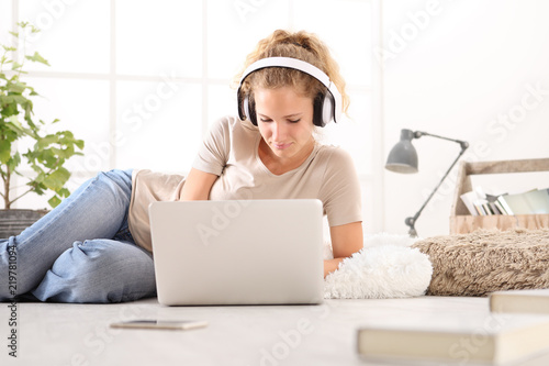 young woman with computer, headphones, smartphone and books, lying on the floor in living room on white wide window in the background