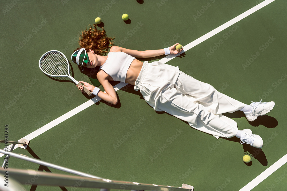 high angle view of stylish woman in white clothing and cap lying with racket lying on tennis court with racket