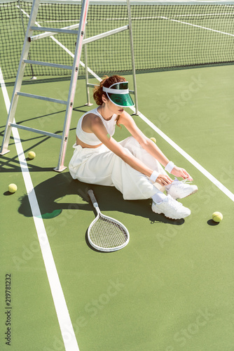 young woman in stylish white clothing and cap tying shoelaces on tennis court with racket and balls © LIGHTFIELD STUDIOS