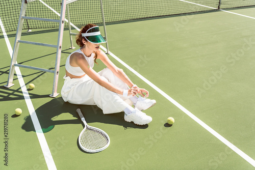 young woman in stylish white clothing and cap tying shoelaces on tennis court with racket and balls © LIGHTFIELD STUDIOS