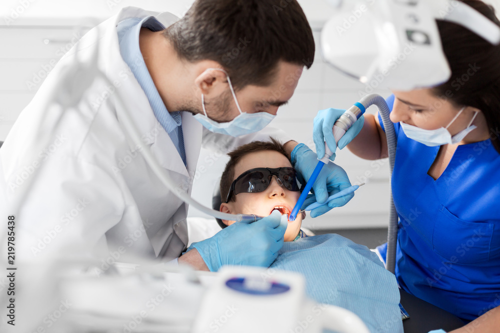 medicine, dentistry and healthcare concept - dentist and assistant with dental drill and saliva ejector treating kid patient teeth at dental clinic medicine, dentistry and healthcare concept - dentist and assistant with dental drill and saliva ejector treating kid patient teeth at dental clinic