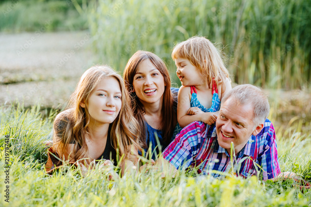 Fototapeta premium Ideal picture of family happiness - happy family with two daughter having fun and lying on the grass in lake shore on a summer day in sunset. Parenthood, love, vacation and happy family concept.