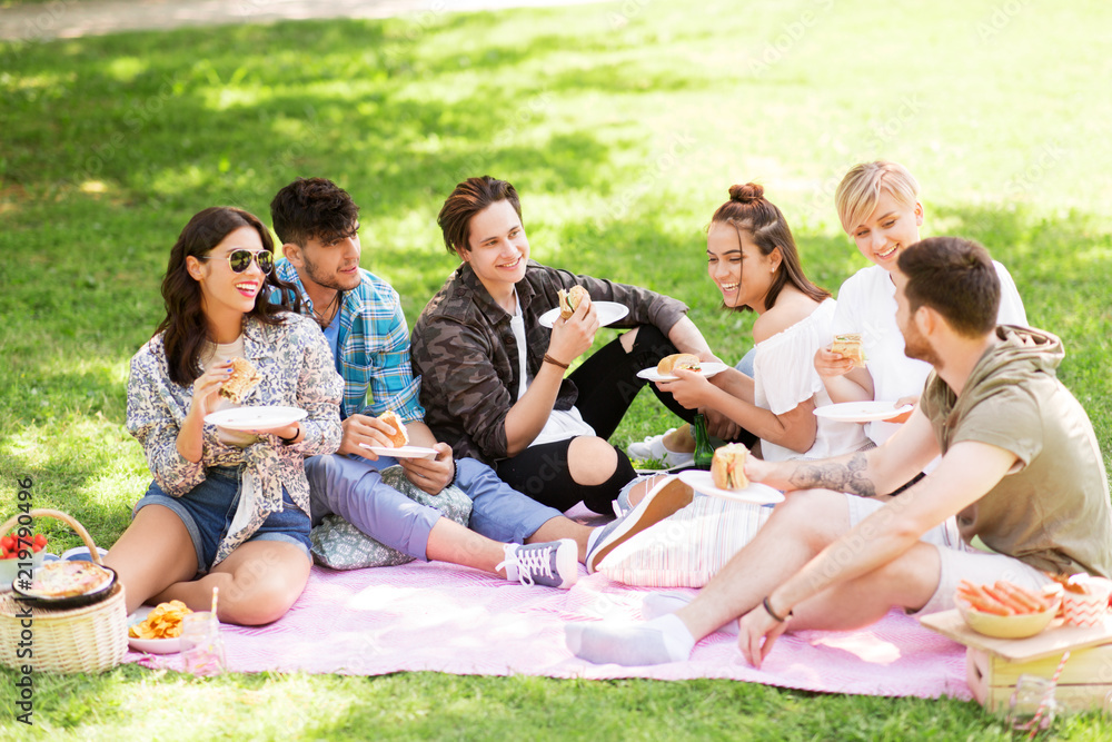 friendship, leisure and fast food concept - group of happy friends eating sandwiches or burgers at picnic in summer park friendship, leisure and fast food concept - group of happy friends eating sandwiches or burgers at picnic in summer park