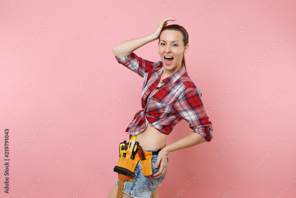 Strong young handyman woman wearing plaid shirt, denim shorts, kit tools belt full of variety useful instruments isolated on pink background. Female doing male work. Renovation and occupation concept.