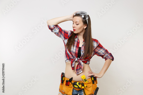 Strong young handyman woman in plaid top shirt, denim shorts, kit tools belt full of instruments in protective goggles isolated on white background. Female in male work. Copy space for advertisement.