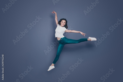 Graceful magnificent attractive pretty stylish trendy nice lovely cheerful curly-haired brunette girl in casual white t-shirt and jeans, flying in air, dancing like ballet, isolated on grey background
