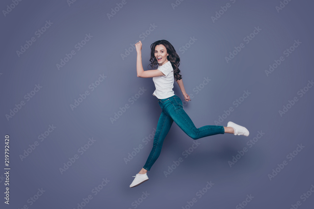 Slim funky attractive adorable pretty stylish trendy nice graceful cheerful curly-haired brunette girl in casual white t-shirt and jeans, running rush hour in air, isolated on grey background