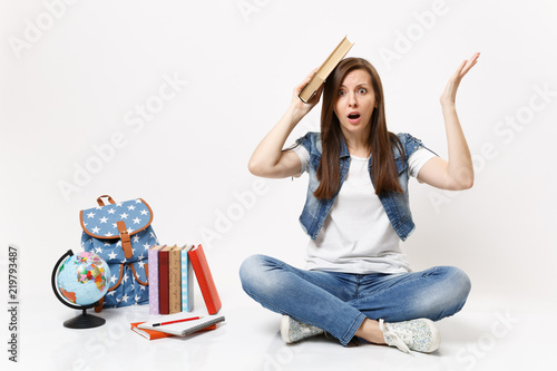 Young shocked woman student in denim clothes holding book near head spreading hand sitting near globe, backpack, school books isolated on white background. Education in high school university college.