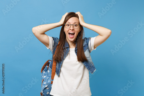 Portrait of young shocked amazed happy woman student in denim clothes glasses with backpack screaming clinging to head isolated on blue background. Education in high school university college concept.