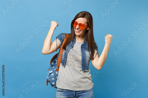 Young woman student with closed eyes with backpack in orange heart glasses clenching fists like winner or happy human isolated on blue background. Education in college. Copy space for advertisement.