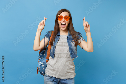 Young excited woman student with opened mouth wearing orange heart glasses waiting for special moment, keeping fingers crossed isolated on blue background. Education in high school. Advertising area.