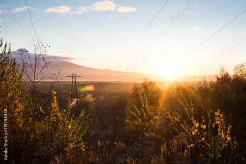 Sunrise is during great morning in mountains. Big metal high-voltage line is in right side. Beautiful grass and trees are under warm light. Sky is highlighted by the sun. Golden autumn in mountains.