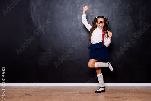 Full size body length of glad smart cute stylish small little girl with curly ponytails in white blouse shirt and blue skirt with red bag, celebrating. Isolated over black background © deagreez
