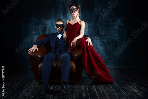 Rich couple man male woman female in love sitting in leather sofa in expensive suit and red evening dress and black carnaval mask. Sex, tempts, harassment, sexism, seduction issues