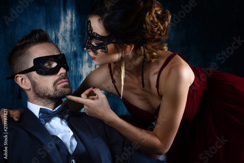 Sexy woman female in black carnaval mask red expensive dress and gold earnings seduces millionaire man male in suit, bow tie and black carnaval mask. Sex, tempts, harassment, sexism, seduction issues photo