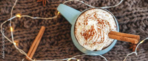 Mug of coffee, cocoa or hot chocolate with whipped cream and cinnamon on scarf with garland, anise star. Pumpkin latte - cozy drink for cold autumn or winter. Flat lay. Top view.