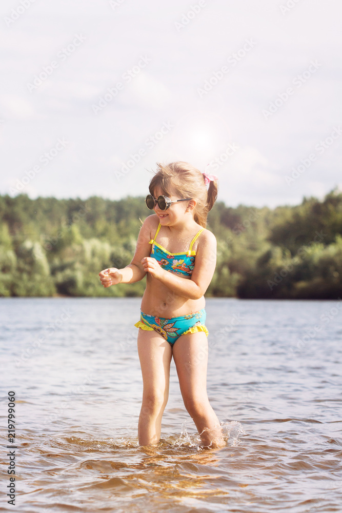 a small blonde girl with a tail in a swimsuit and glasses fooling around jumping splashes in the sunny warm summer weather in the river