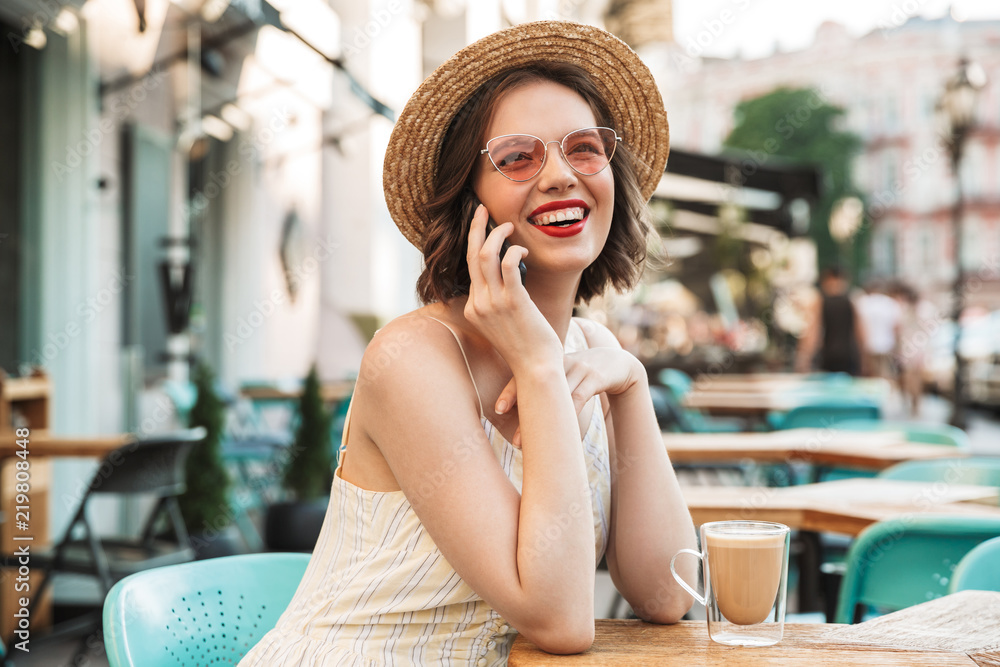 Joyful woman in dress and straw hat talking by smartphone