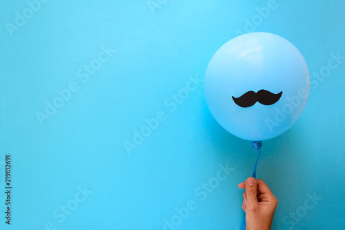 Hand holding blue balloon with a paper mustache on blue paper background. Cut out style. Movember concept. Top view. Flat lay. Copy space photo