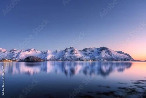 Obraz Piękny śnieg pokryte górami i kolorowe niebo odbite w wodzie w nocy. Zimowy pejzaż z morzem, zaśnieżone skały, błękitne niebo, odbicie, światło słoneczne o zachodzie słońca. Lofoty, Norwegia o zmierzchu. Natura