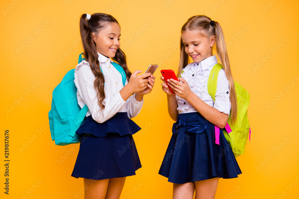 Profile side view photo of two happy and focused small girls in skirts isolated on bright yellow background with smartphones in their hands read interesting news Profile side view photo of two happy and focused small girls in skirts isolated on bright yellow background with smartphones in their hands read interesting news