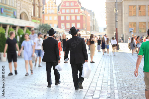 Dwóch mężczyzn w czarnych kapeluszach i garniturach idą z reklamówkami przez miasto.