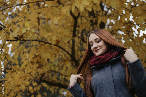 The girl touching hair. Young romantic female person wearing warm grey overcoat and red scarf. Model standing in autumn park on the tree with yellow leaves background.