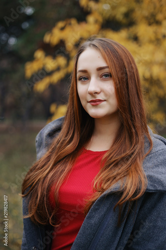Young romantic female person wearing warm grey overcoat. Model standing in autumn park on the tree with yellow leaves background. Pretty girl looking at camera with brooding look.