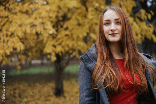 Young romantic female person wearing warm grey overcoat. Model standing in autumn park on the tree with yellow leaves background. Pretty girl looking at camera with brooding look.