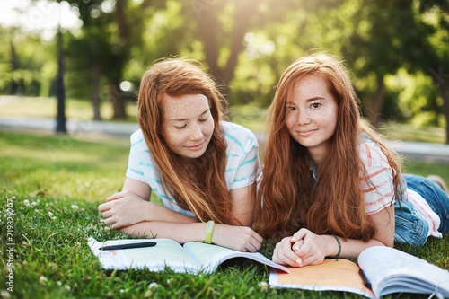 Friendship, family and lifestyle concept. Two attractive redhead girls relaxing on sunny day, lying on grass and doing homework together, smiling at camera with carefree expression