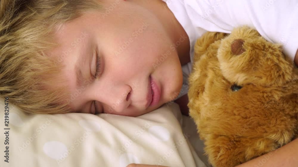 Closeup portrait of cute funny young white kid lying in his bed with cute brown teddy bear toy and pretending to be sleeping. Then suddenly looks at camera smiling. 4k video footage. Closeup portrait of cute funny young white kid lying in his bed with cute brown teddy bear toy and pretending to be sleeping. Then suddenly looks at camera smiling. 4k video footage.
