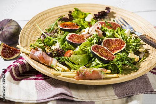 gemischter Blattsalat mit Feigen, Tomaten, Schafskäse und Crossinis mit Schinken,serviert auf Holzteller, weisser Holztisch 