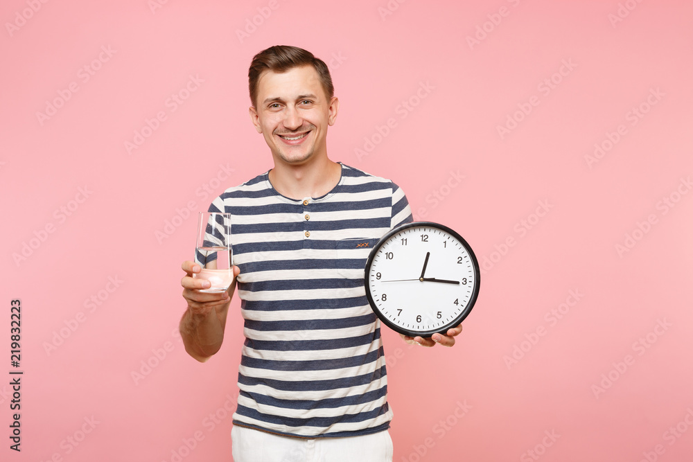 Portrait of happy caucasian man in striped t-shirt holding round clock, clear fresh pure water in glass isolated on trending pastel pink background. People healthy lifestyle concept. Time running out. Portrait of happy caucasian man in striped t-shirt holding round clock, clear fresh pure water in glass isolated on trending pastel pink background. People healthy lifestyle concept. Time running out.