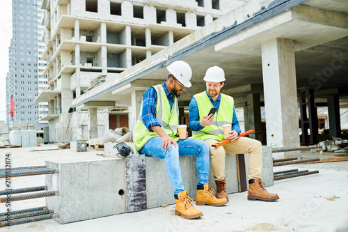 Full length portrait of two workers wearing hardhats  chatting and using smartphone during coffee break on construction site, copy space photo