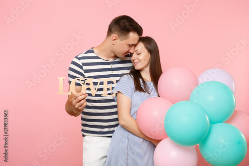 Tender couple hold wooden word letters love. Woman and man in blue clothes celebrating birthday holiday party on pastel pink background with colorful air balloons. People sincere emotions concept.