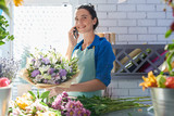 Young smiling florist holding flowers and connecting on mobile phone near workplace