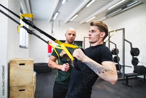 Fit young man with a personal trainer in gym doing exercise with TRX.