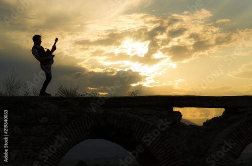 Man performs on electric guitar on old Roman aqueduct during sunset.