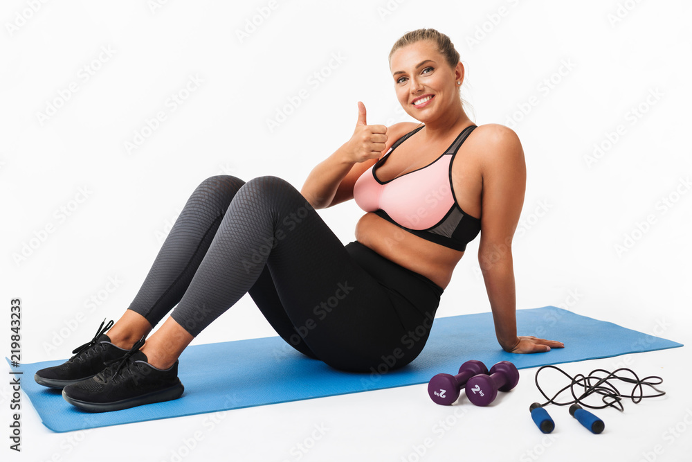 Pretty smiling girl with excess weight in sporty top and leggings sitting  on yoga mat while happily showing thumb up gesture looking in camera over  white background. Plus size model Stock Photo