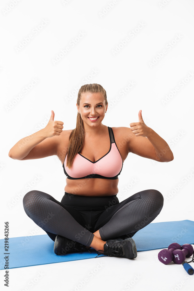 Pretty girl with excess weight in sporty top and leggings sitting on yoga mat joyfully showing big thumbs up looking in camera over white background isolated. Plus size model Pretty girl with excess weight in sporty top and leggings sitting on yoga mat joyfully showing big thumbs up looking in camera over white background isolated. Plus size model
