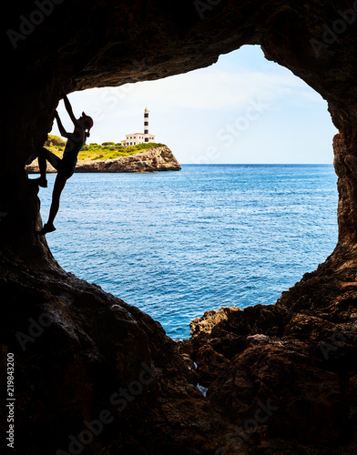 Silhouette of a female athlete climbing in a cave, Portocolom Lighthouse on a cliff in background, Mallorca, Spain.