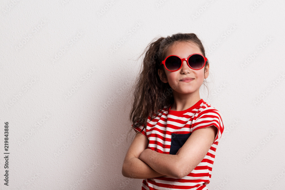A small girl with red sunglasses and striped T-shirt standing in a studio. A small girl with red sunglasses and striped T-shirt standing in a studio.