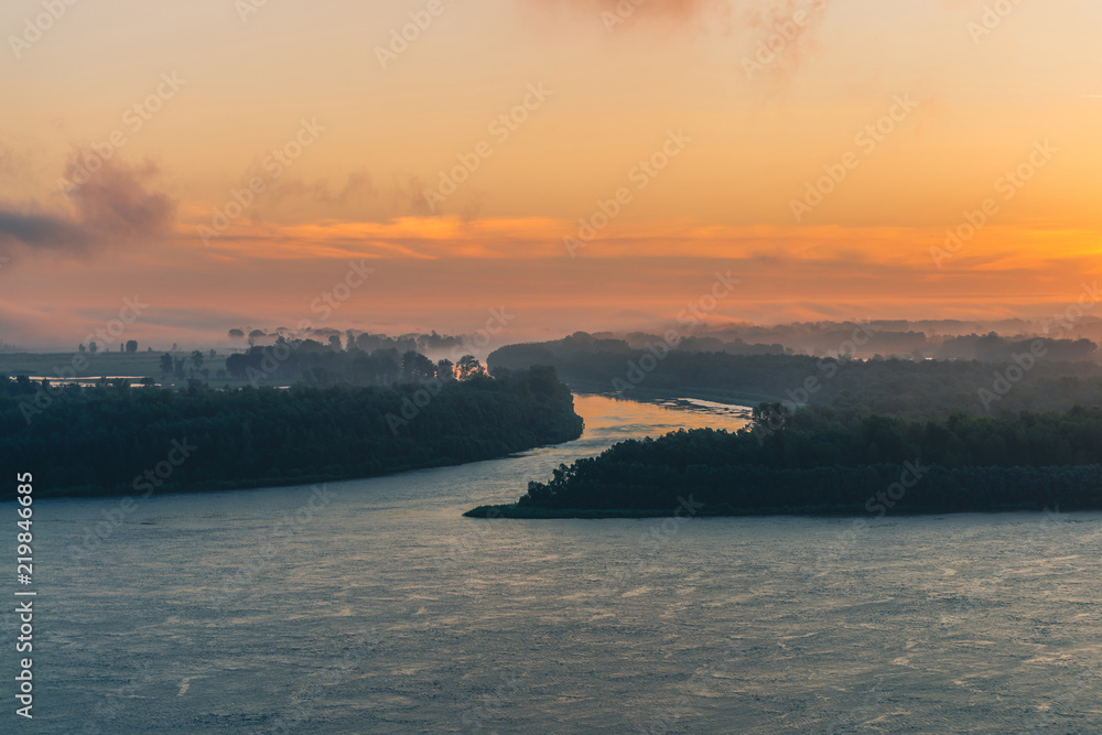 Broad river flows along shore with forest under mist. Channel of river flows around island. Orange glow at dawn reflected on water. Colorful morning mystical atmospheric landscape of majestic nature.