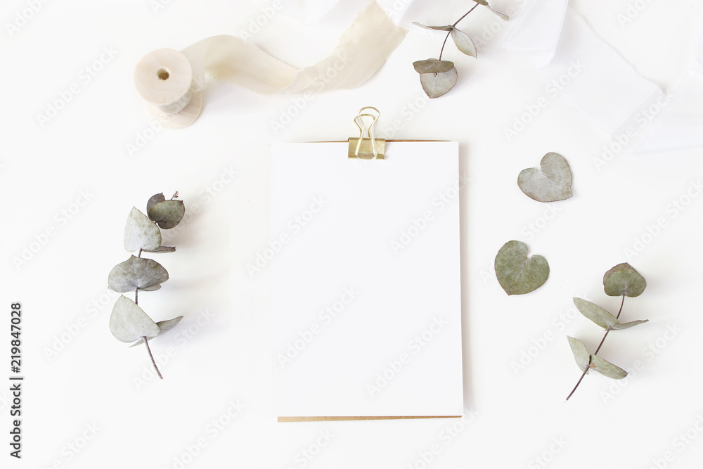 Feminine wedding desktop stationery mockup with blank greeting card, dry eucalyptus leaves, silk ribbon and golden binder clip on white table background. Flat lay, top view. Styled stock photo. - obrazy, fototapety, plakaty 