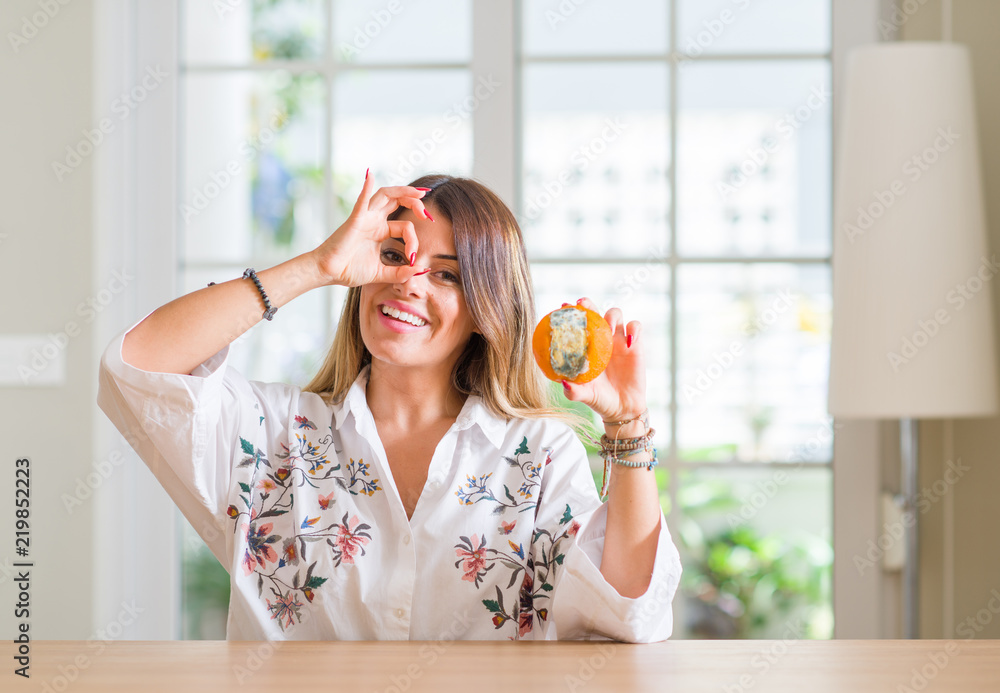 Young woman at home holding rotten orange with happy face smiling doing ok sign with hand on eye looking through fingers Young woman at home holding rotten orange with happy face smiling doing ok sign with hand on eye looking through fingers