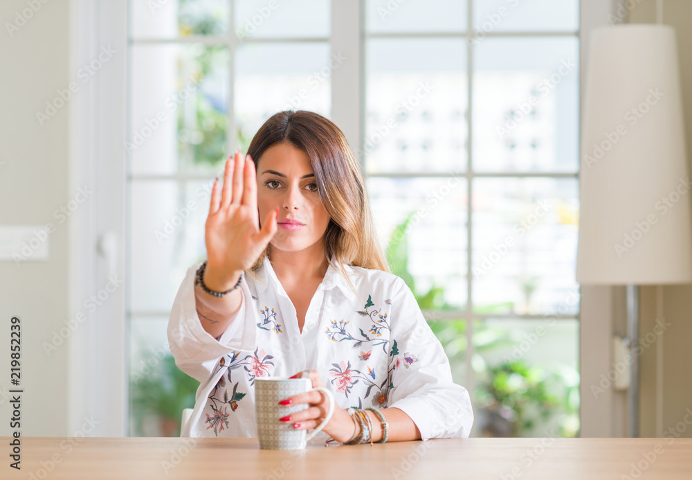 Young woman at home drinking coffee with open hand doing stop sign with serious and confident expression, defense gesture Young woman at home drinking coffee with open hand doing stop sign with serious and confident expression, defense gesture