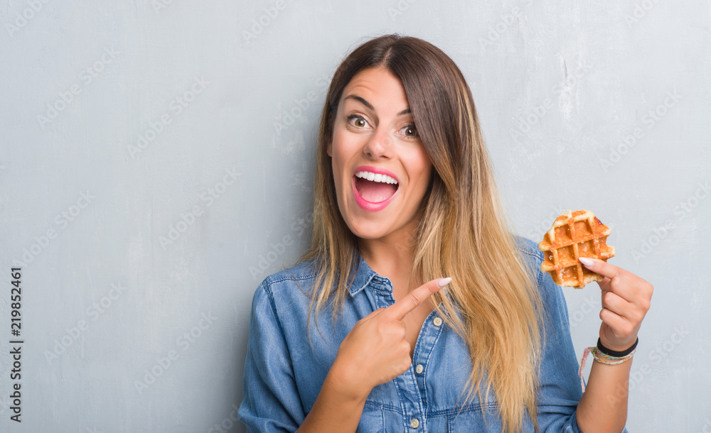 Young adult woman over grey grunge wall eating Belgium waffle very happy pointing with hand and finger Young adult woman over grey grunge wall eating Belgium waffle very happy pointing with hand and finger