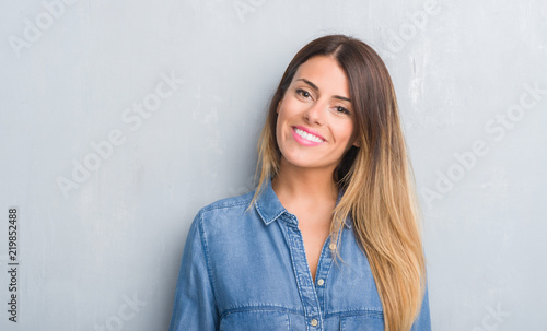 Young adult woman over grey grunge wall wearing denim outfit with a happy face standing and smiling with a confident smile showing teeth photo