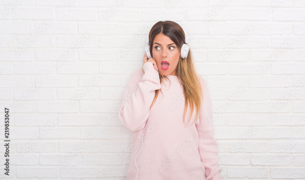 Young adult woman over white brick wall wearing headphones listening to music and dancing scared in shock with a surprise face, afraid and excited with fear expression