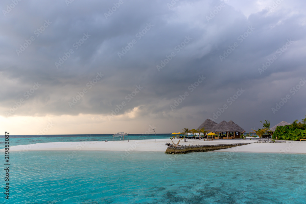 An idyllic island in Maldive in a stormy weather afternoon. Super clear water, white sand, traditional huts and tropical climate feeling.